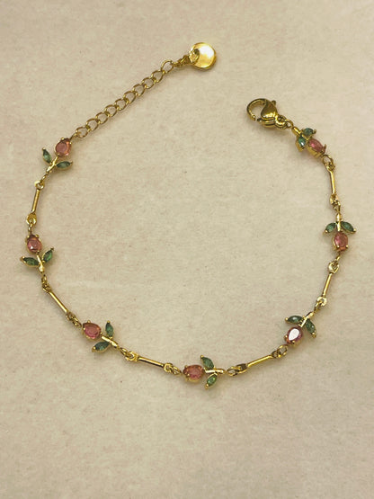 A gold chain bracelet featuring small rose motifs with pink faceted stones and green enamel leaves, displayed on a neutral beige background.