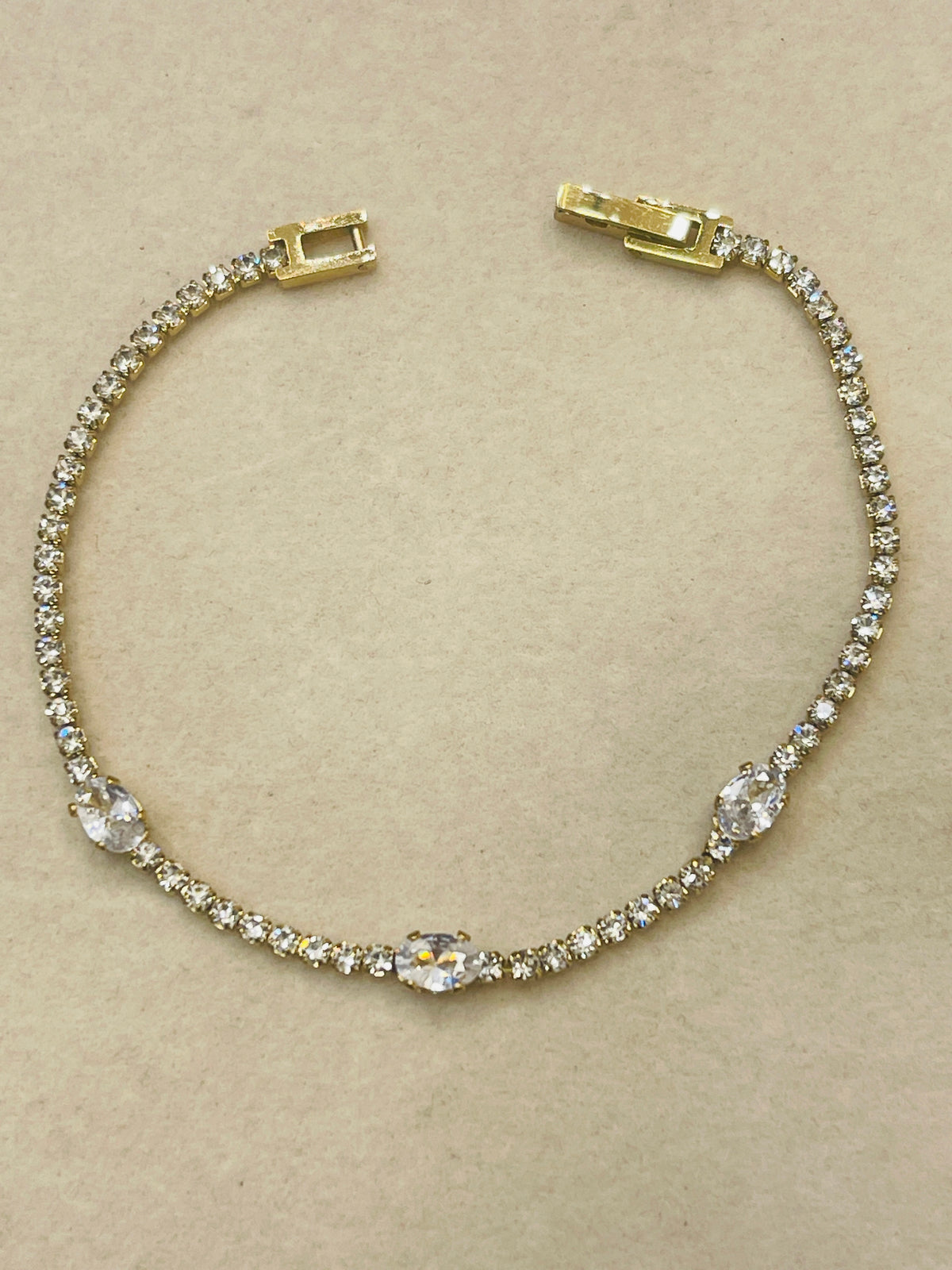 A gold tennis bracelet adorned with clear crystals, featuring a central oval crystal and three smaller matching crystals spaced along the band, displayed on a neutral beige background.