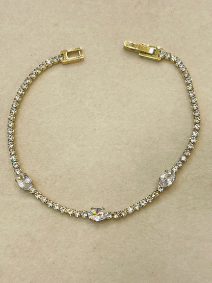 A gold tennis bracelet adorned with clear crystals, featuring a central oval crystal and three smaller matching crystals spaced along the band, displayed on a neutral beige background.