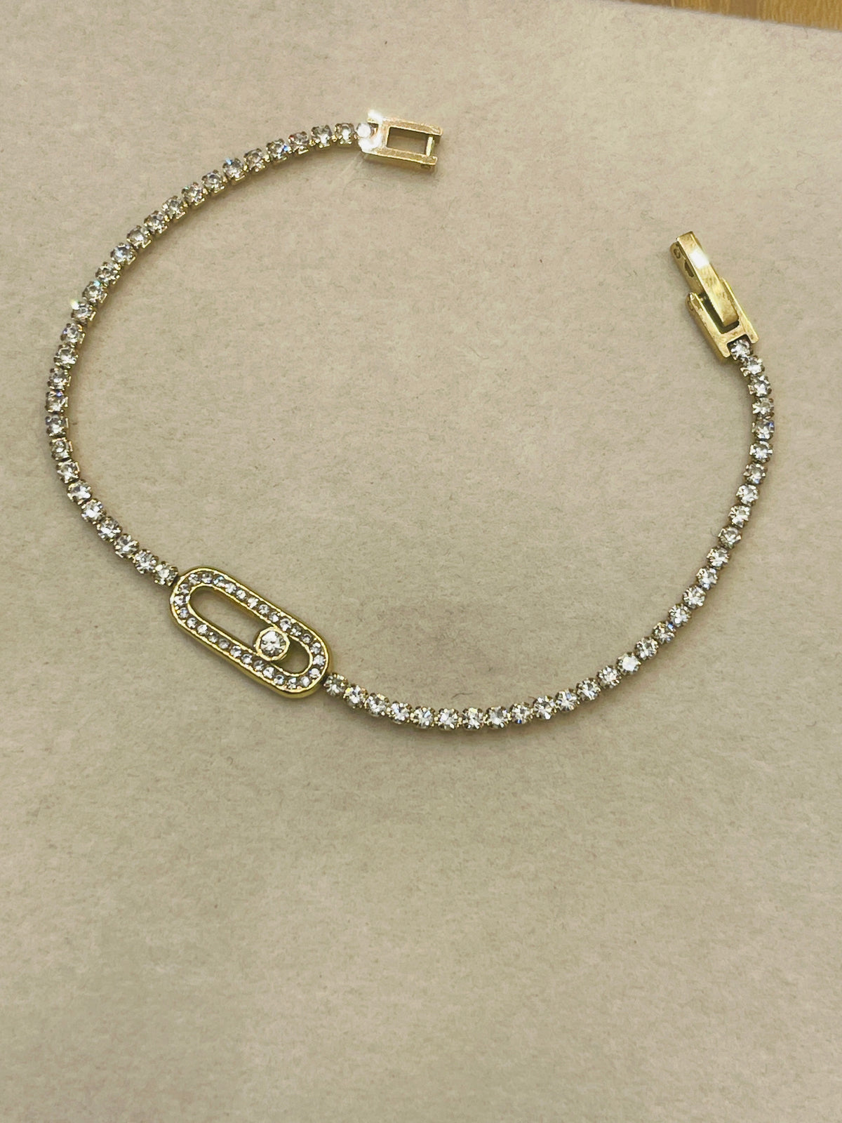 A gold-tone tennis bracelet with clear pavé stones, featuring a central oval link with a small round stone and a lobster clasp closure, displayed on a neutral beige textured surface.