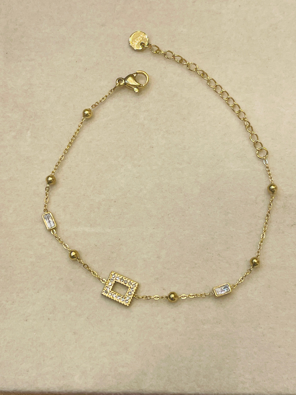 A delicate gold chain bracelet with a central square pavé charm, small gold beads, and rectangular clear crystal accents, displayed on a textured beige surface.