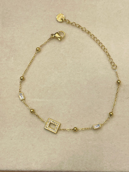 A delicate gold chain bracelet with a central square pavé charm, small gold beads, and rectangular clear crystal accents, displayed on a textured beige surface.