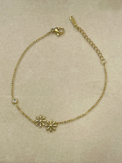 A delicate gold chain bracelet with two small starburst charms and a single clear rhinestone accent, displayed on a textured beige background.