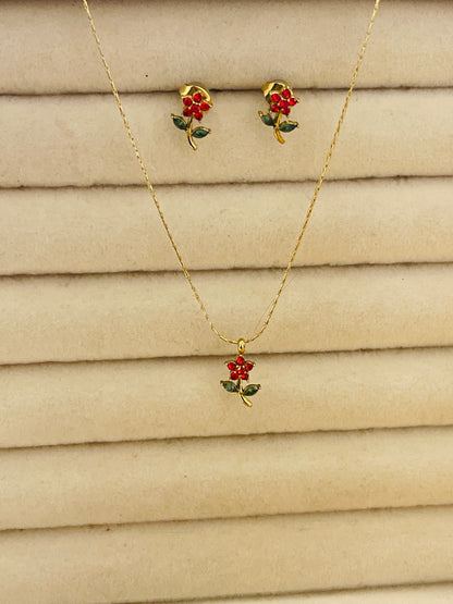 A gold necklace and matching stud earrings set with small red flower and green leaf charms, displayed on a beige textured background.