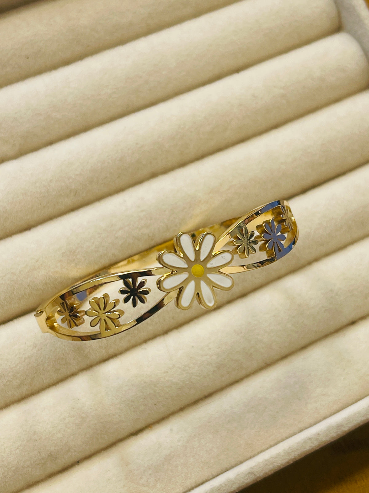 A gold filigree bangle bracelet with white enamel flower details, displayed on a beige jewelry box.
