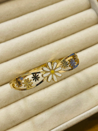 A gold filigree bangle bracelet with white enamel flower details, displayed on a beige jewelry box.