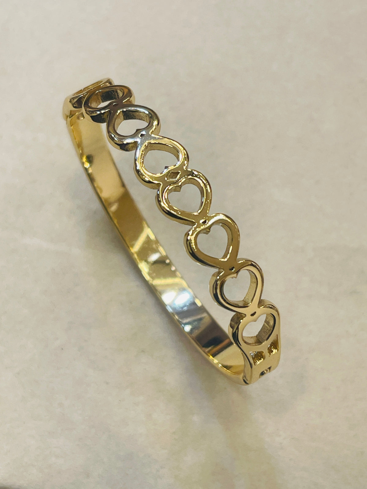 A gold bangle bracelet featuring a row of interlocking open heart shapes, displayed on a neutral beige background.