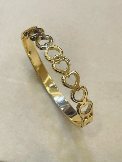 A gold bangle bracelet featuring a row of interlocking open heart shapes, displayed on a neutral beige background.