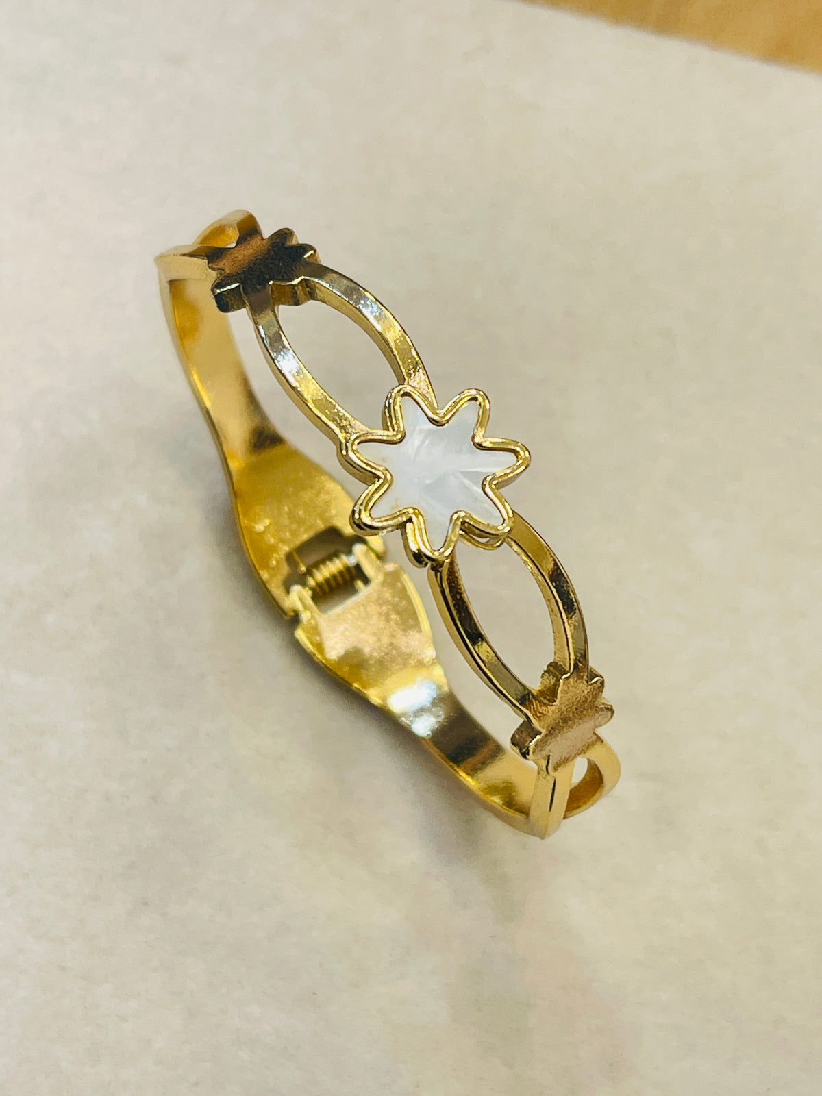 A gold bangle bracelet featuring a central white mother-of-pearl star-shaped inlay and small gold leaf accents, displayed on a light beige textured surface.