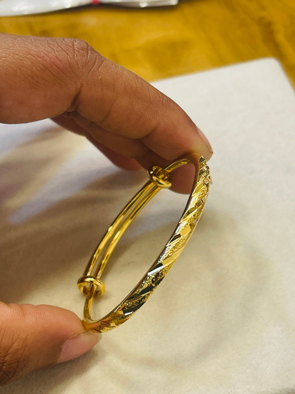 A close-up of a hand holding a gold-plated bangle bracelet with a textured, twisted diamond pattern on a white surface.
