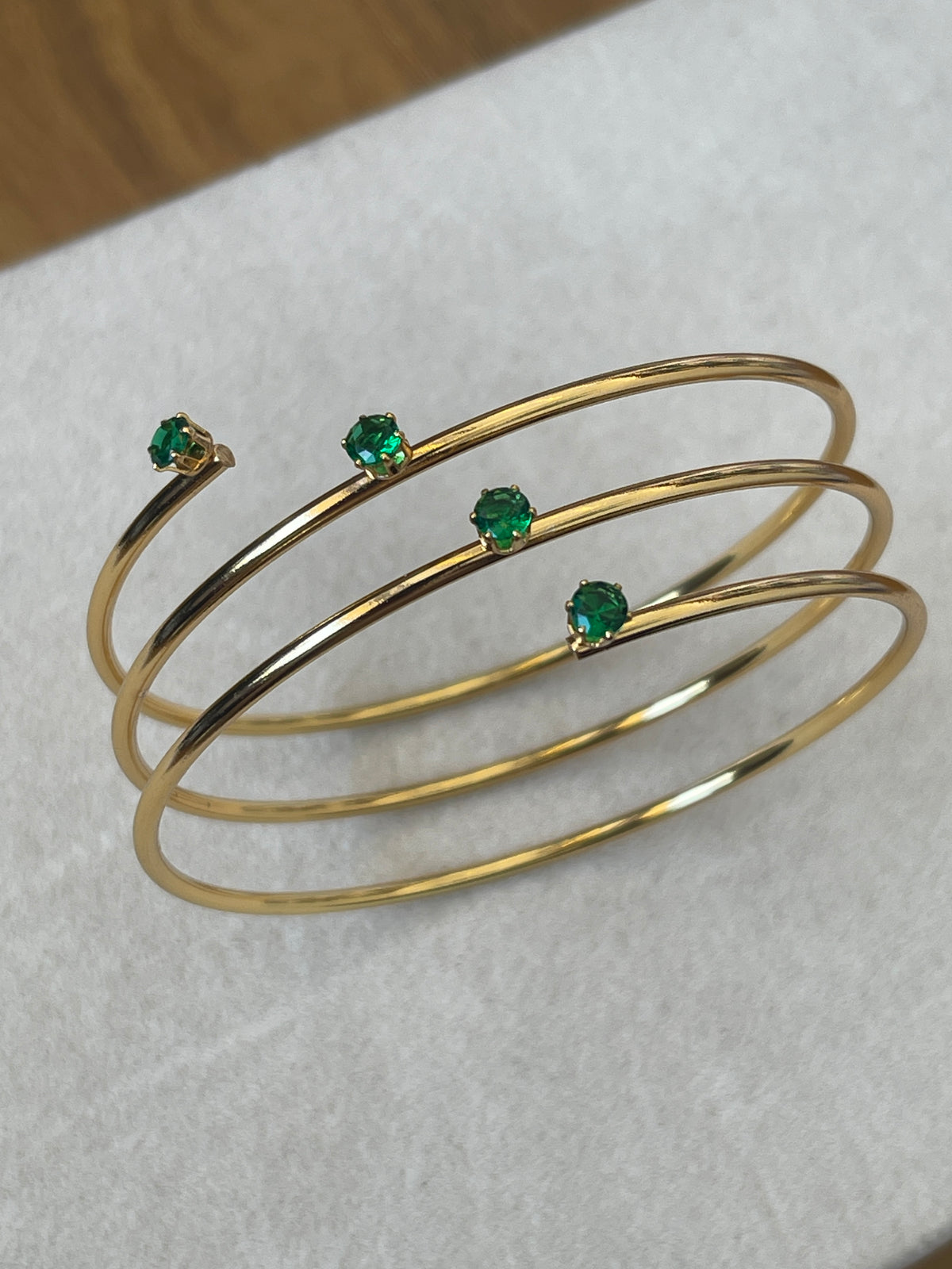 Three thin gold bangle bracelets with small round green gemstone accents, displayed on a light textured surface.