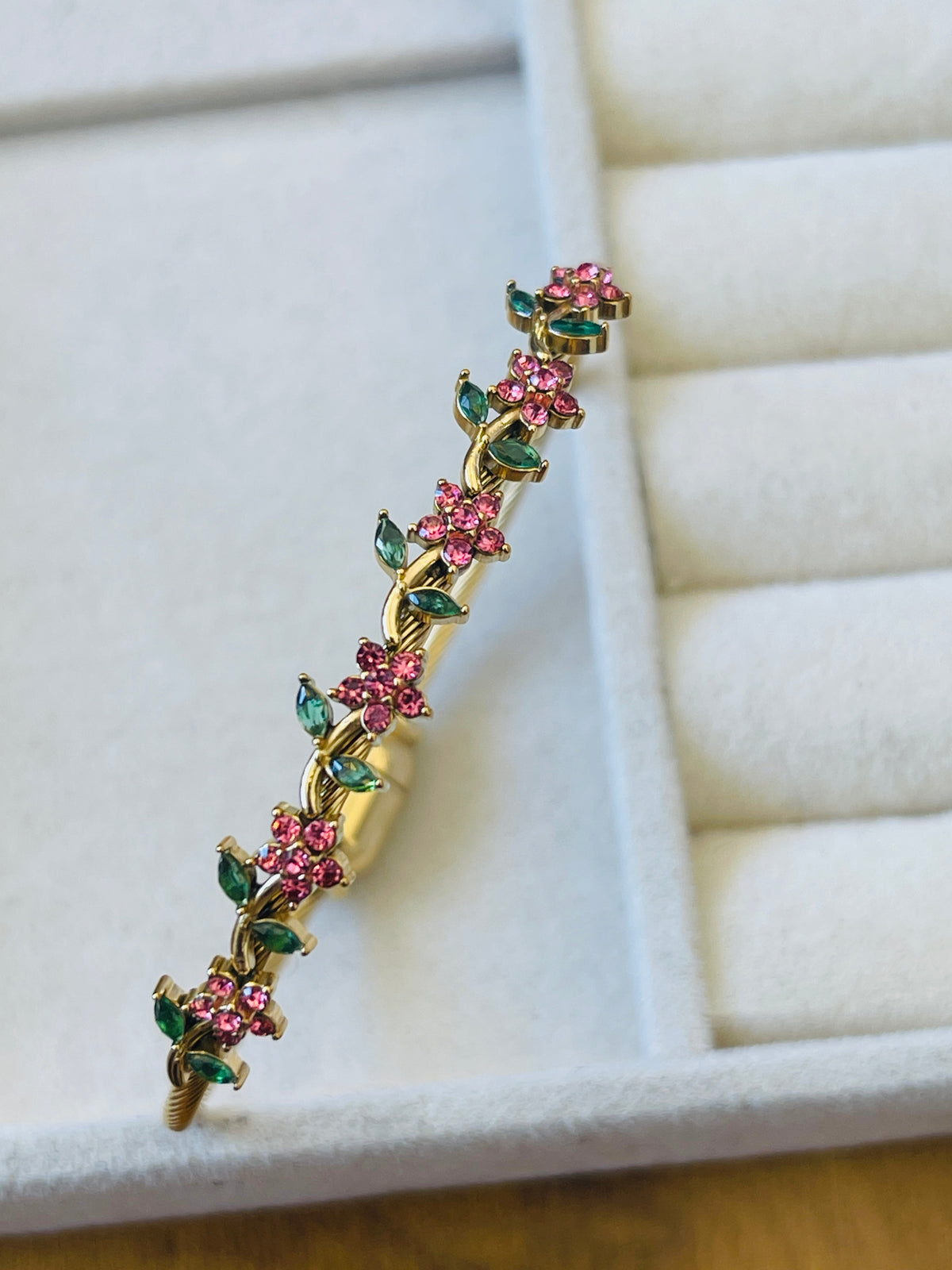A gold-toned floral hair claw clip decorated with pink gemstone flowers and green leaf accents, displayed on a cream-colored jewelry box.