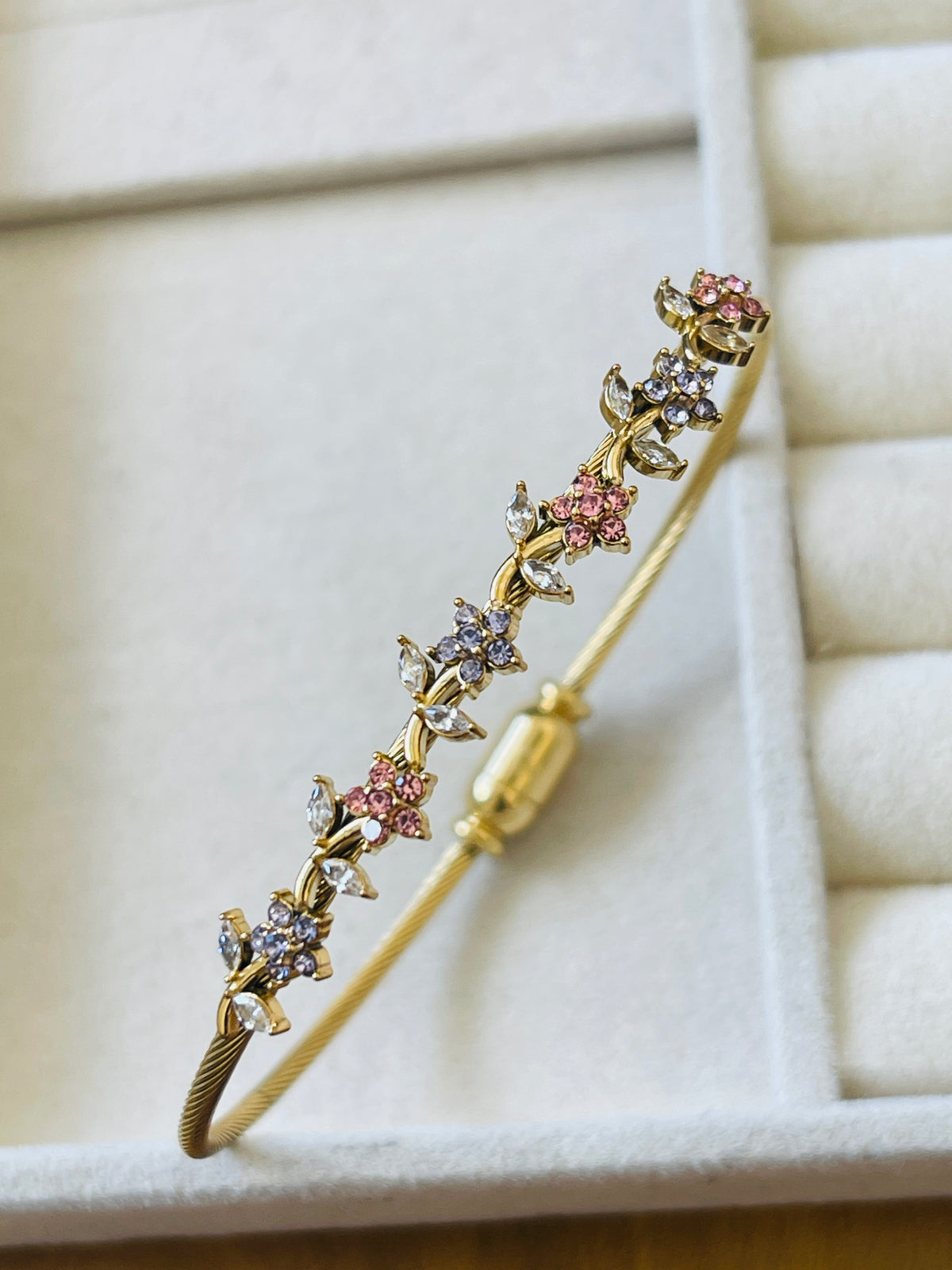 A gold bangle bracelet decorated with small floral motifs set with pink and clear stones, displayed on a cream-colored jewelry box.