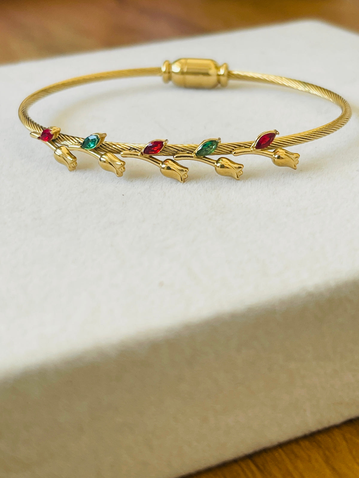 A gold bangle bracelet with small gold tulip-like charms and red and green jewel accents, displayed on a cream-colored jewelry box.