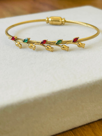 A gold bangle bracelet with small gold tulip-like charms and red and green jewel accents, displayed on a cream-colored jewelry box.