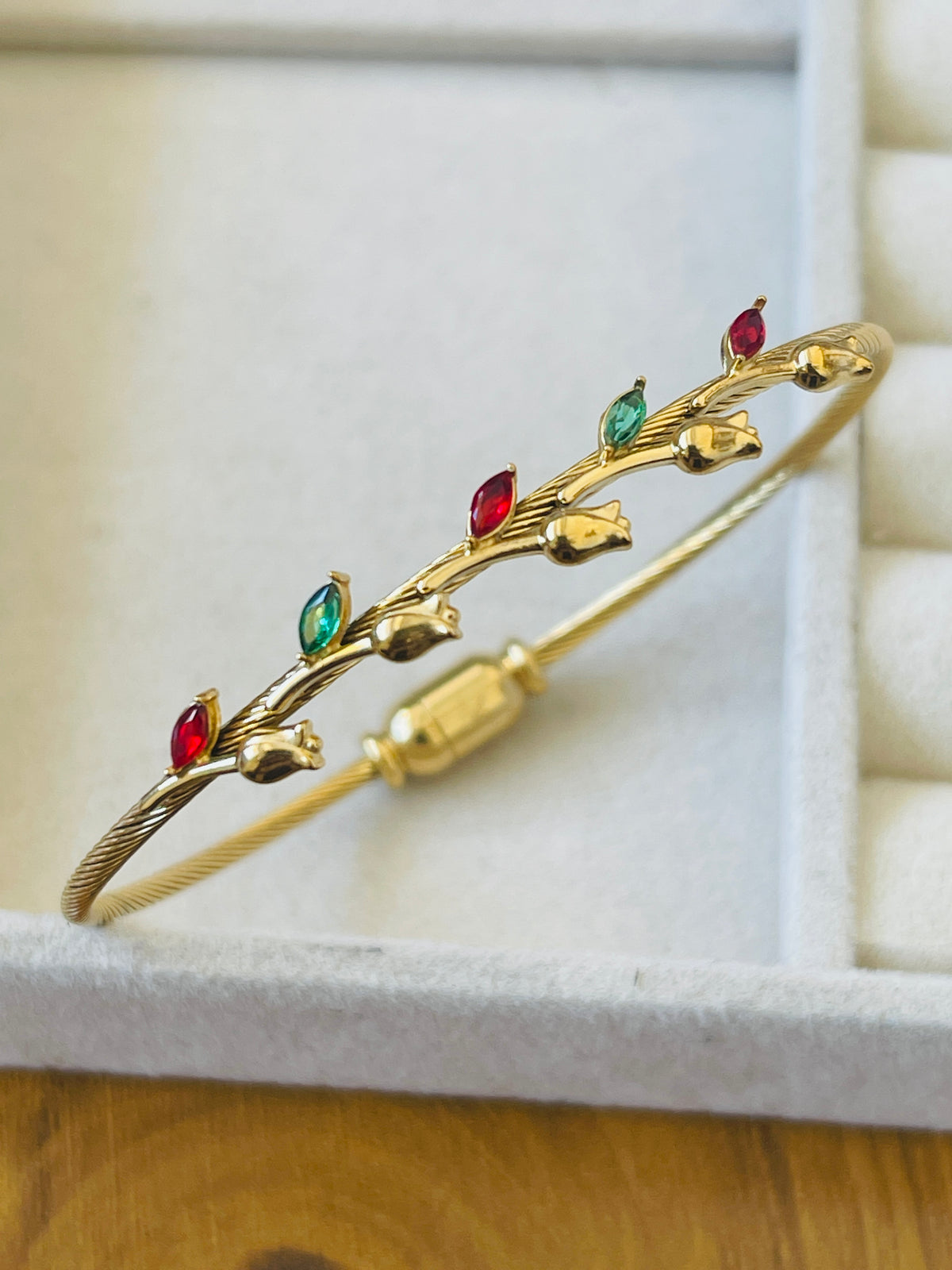 A gold-plated twisted wire bangle bracelet decorated with small gold leaf motifs and marquis-cut red and green gemstones, resting on a white jewelry box.