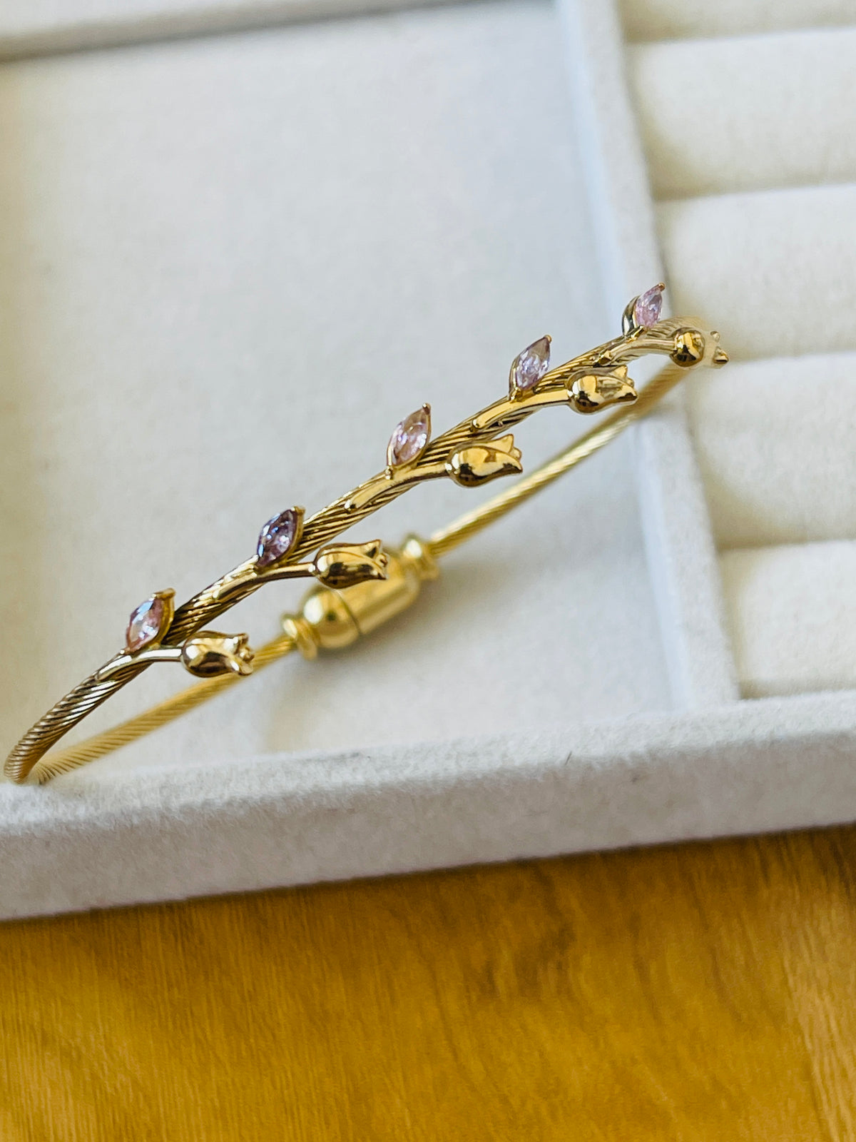 A gold bangle bracelet with a twisted branch design, featuring small gold leaves and marquise-cut pink gemstones, displayed on a white jewelry box.