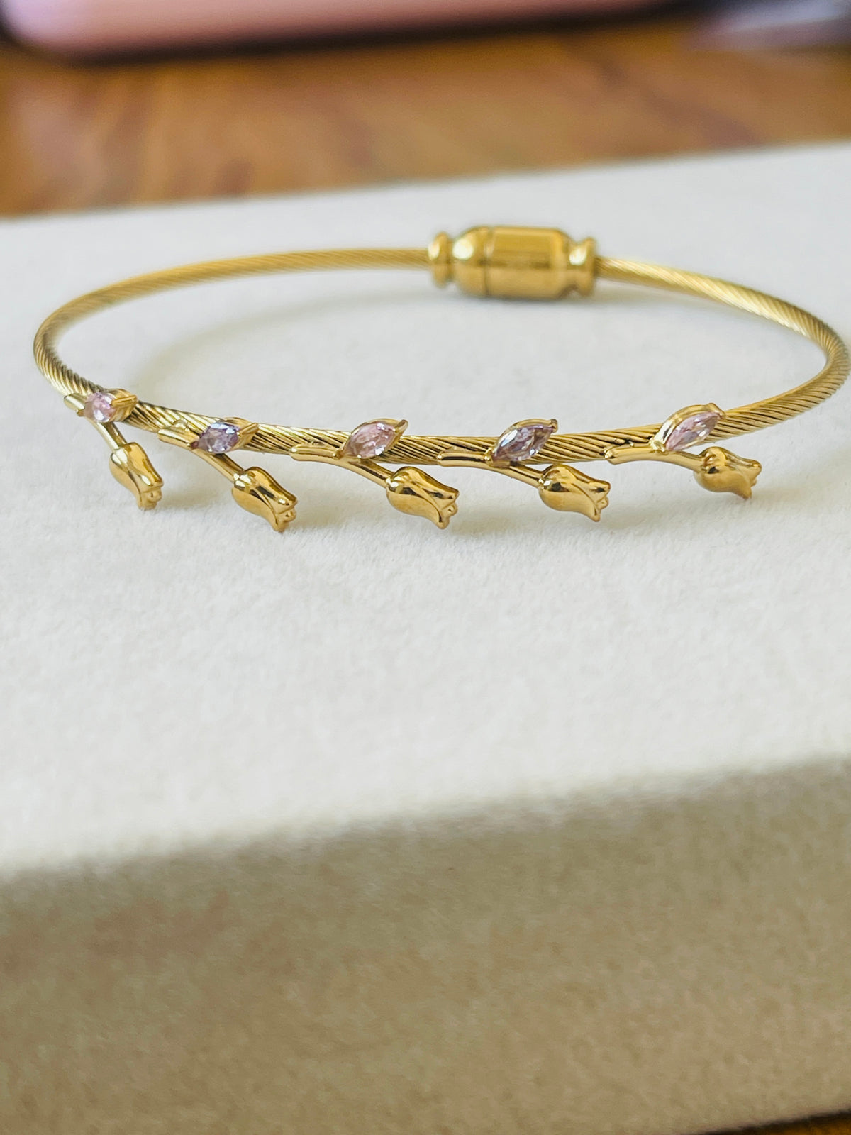 A gold-plated twisted wire bangle bracelet with small gold flower buds and pink marquise-cut gemstones, resting on a textured cream-colored jewelry box.