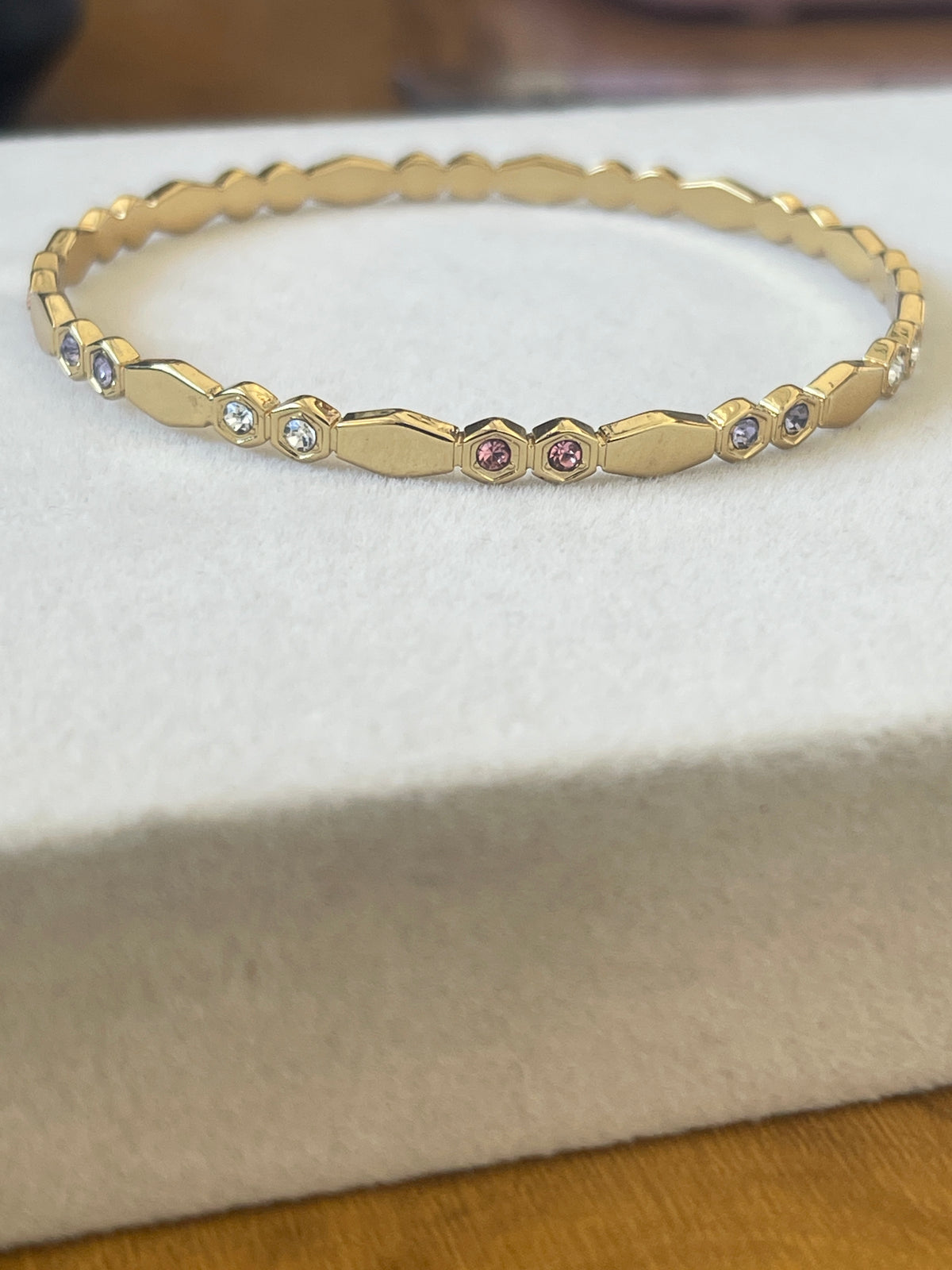 A gold bangle bracelet with a textured geometric pattern and small inset gemstones, displayed on a white textured jewelry box.
