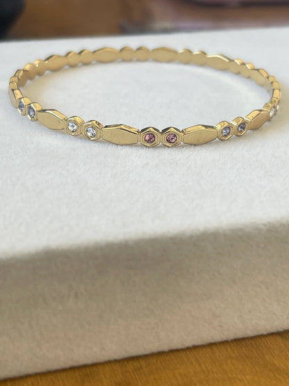 A gold bangle bracelet with a textured geometric pattern and small inset gemstones, displayed on a white textured jewelry box.