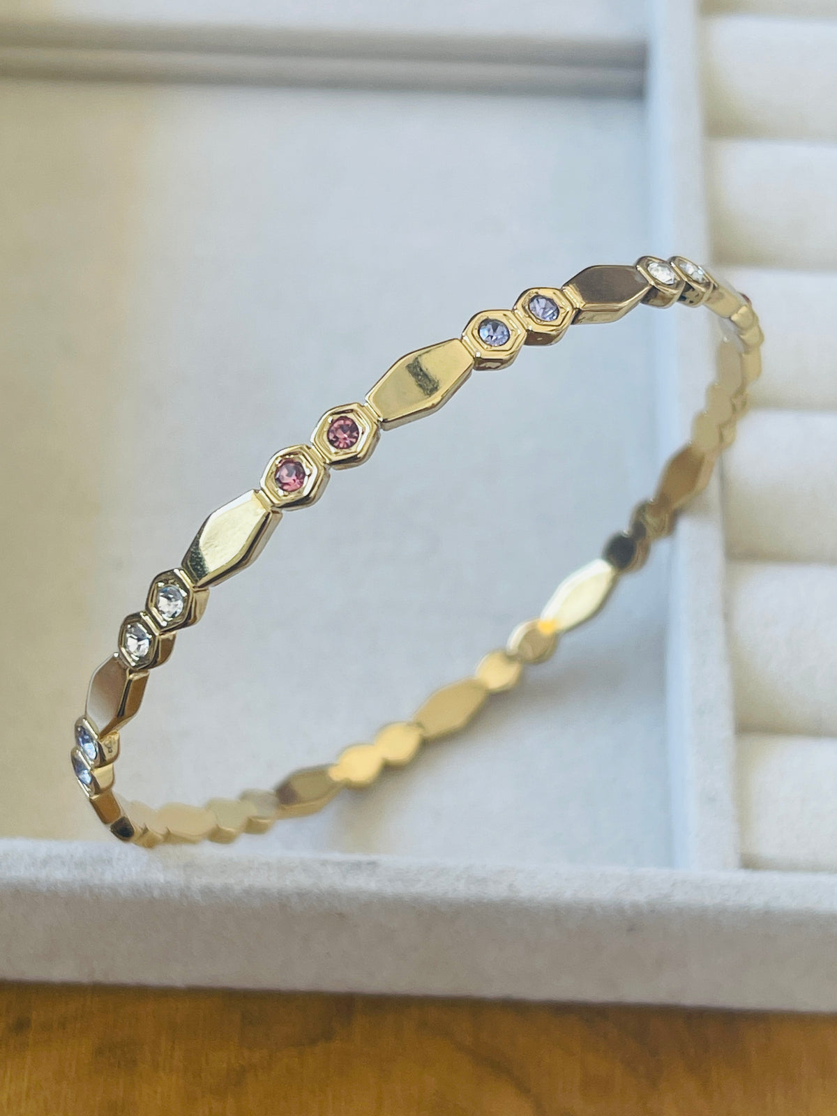 A gold bangle bracelet with a textured geometric pattern and small inset stones, displayed on a white jewelry box.
