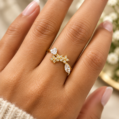 A close-up of a hand wearing a gold ring with clear gemstones in a floral cluster design, set against a soft, blurred background.