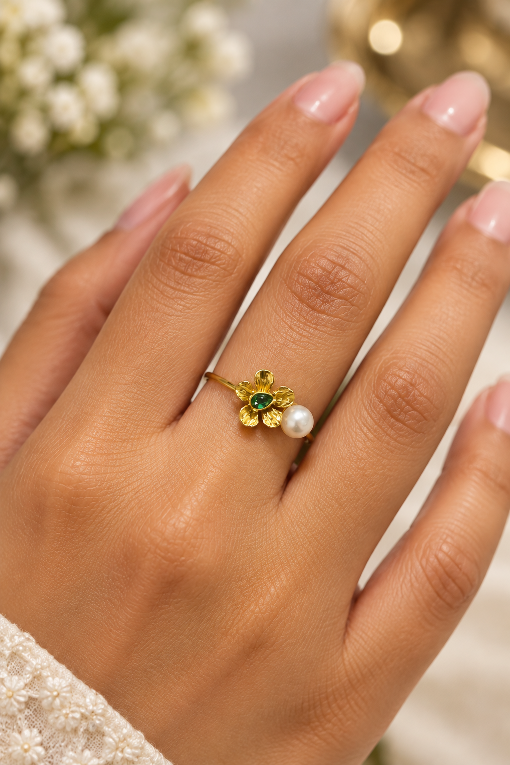 A gold ring with a flower design, featuring a green emerald-cut center and a white pearl, shown worn on a hand with manicured nails.