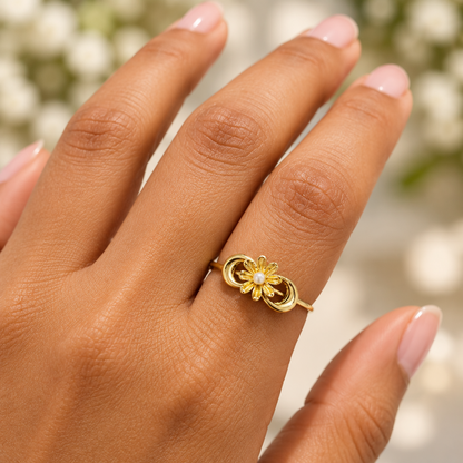 A close-up of a hand wearing a delicate gold ring with a small flower and pearl center, set against a soft, blurred background of white flowers.
