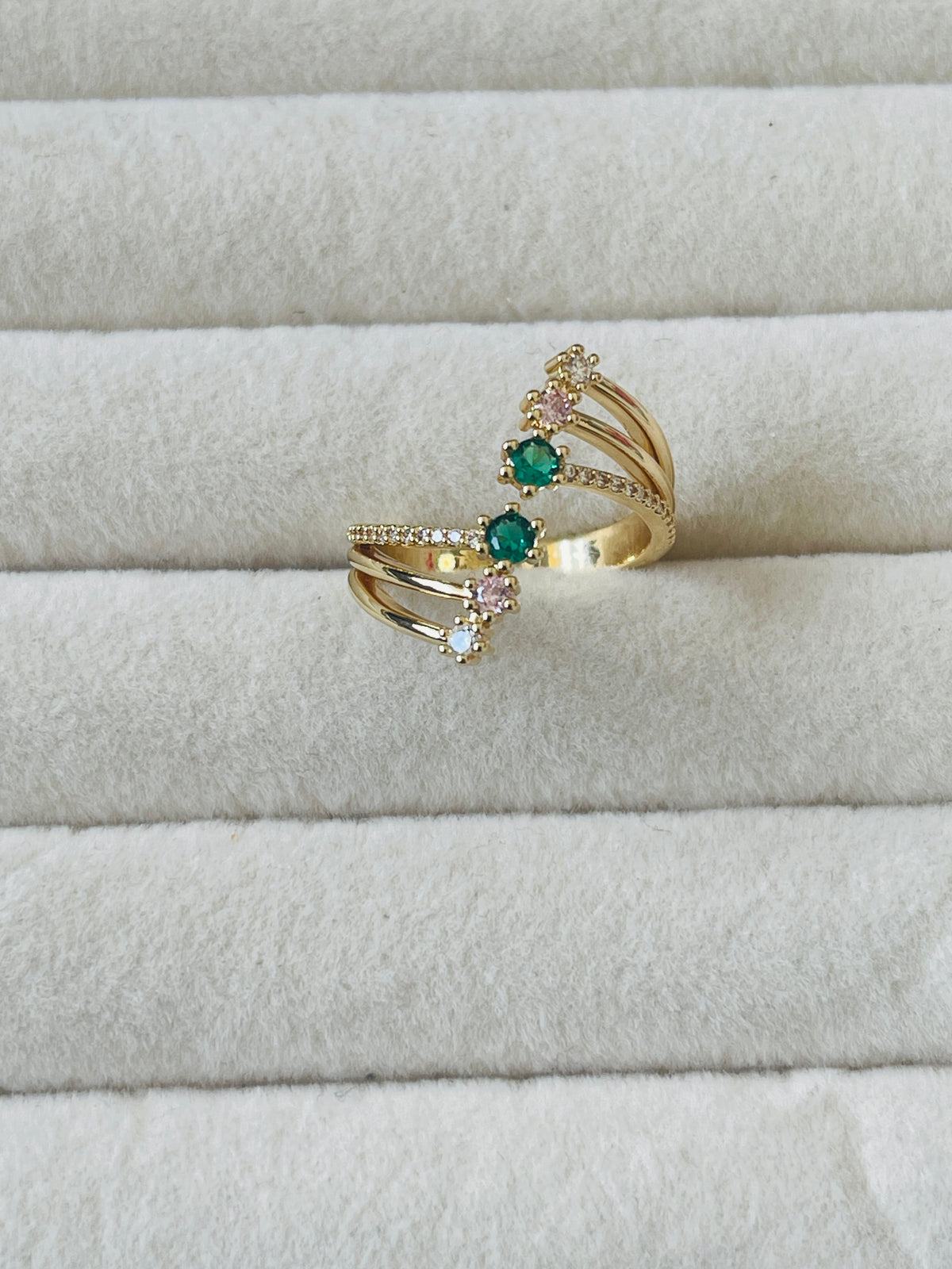 A gold-plated double ring with green and pink gemstone accents, displayed on a textured white ring box.