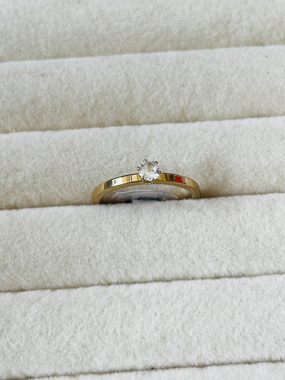 A yellow gold ring with a single round-cut clear gemstone, displayed in the center of a cream-colored velvet ring box.