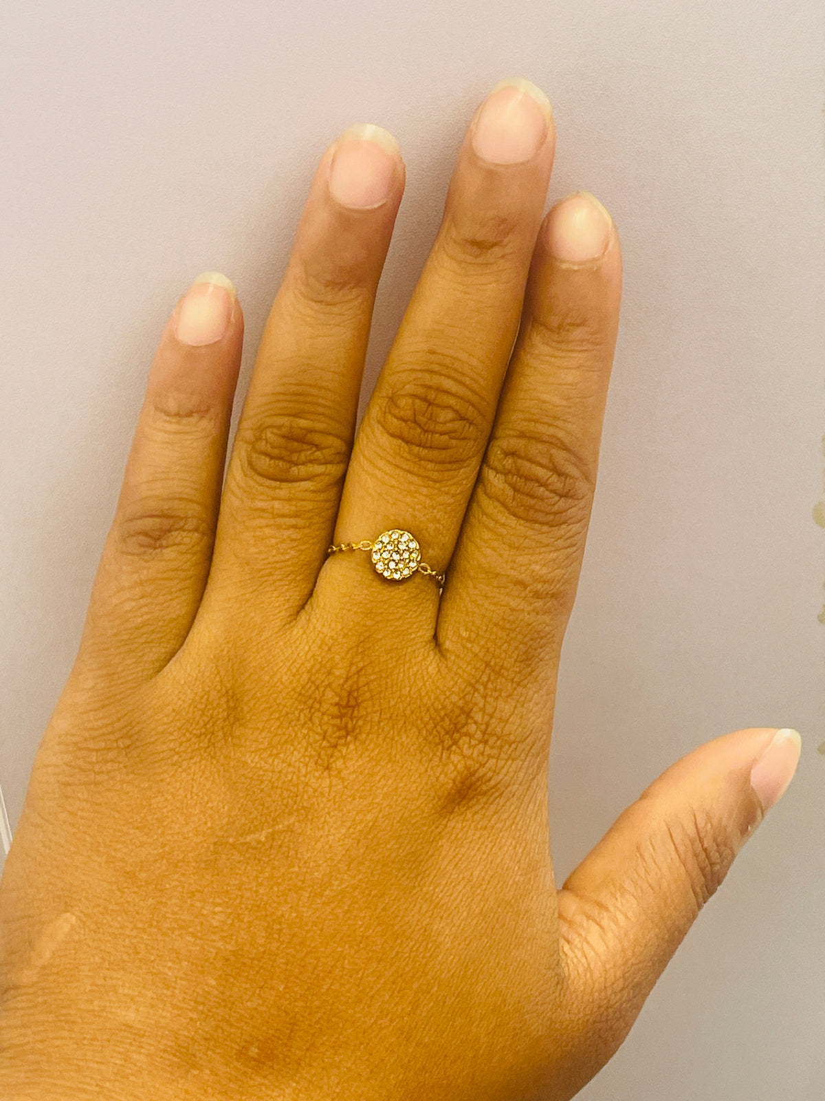 A close-up of a hand wearing a delicate gold ring with a small, round, clear-stone cluster setting on the ring finger, set against a plain light background.