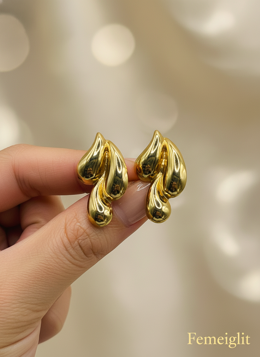 Gold leaf-shaped earrings on a white surface with a white flower in the background