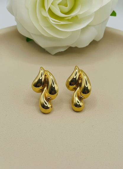 Gold leaf-shaped earrings on a white surface with a white flower in the background