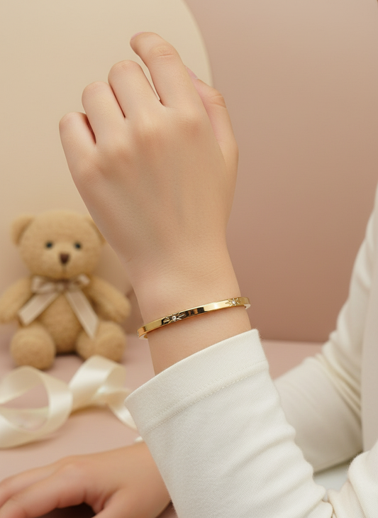 Gold bracelet on a wrist with a teddy bear and ribbon in the background