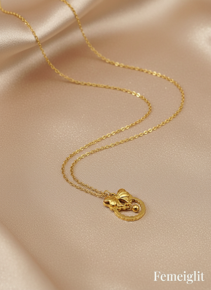Gold necklace with a heart-shaped pendant on a beige fabric background, featuring the brand 'Femeiglit'.