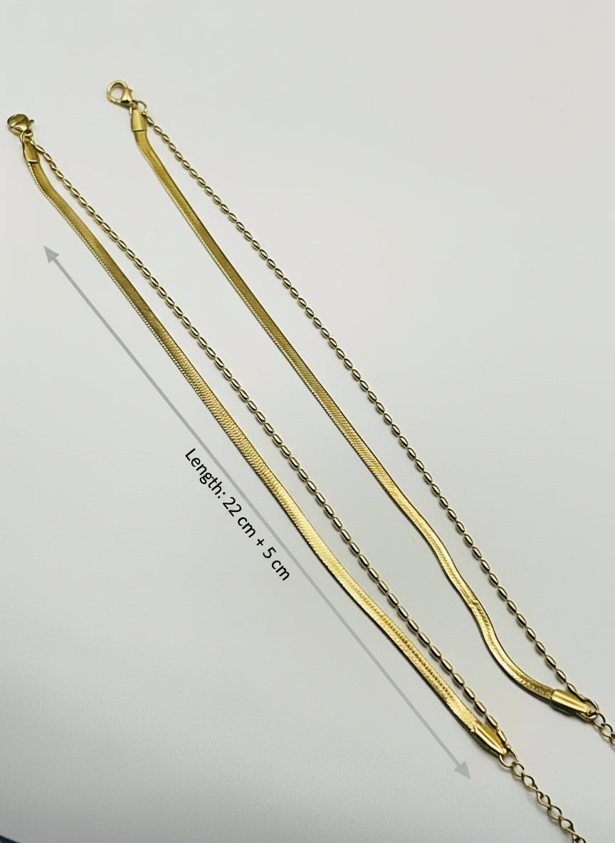 Two gold anklet on a light gray surface with a wooden block in the background.